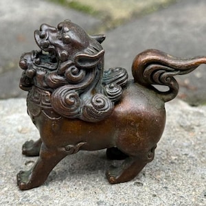 May include: A bronze-colored, detailed statue of a mythical creature, possibly a guardian lion. The sculpture features intricate curls in the mane and tail, with a fierce expression. The statue is on all fours, with a detailed body and paws.