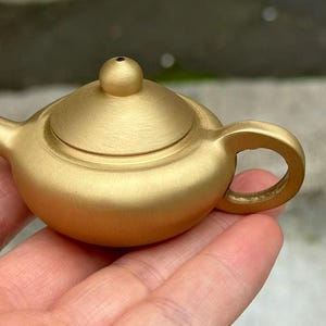 Peut inclure: Une petite théière de couleur or mat avec un corps arrondi, un bec court, une anse incurvée et un couvercle doté d'un petit bouton sphérique. La théière présente une finition métallique lisse et un design circulaire compact.