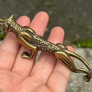 以下が含まれることがあります： 細長く伸びたネコ科の捕食動物の形をした、メタリックゴールド調の真鍮製フィギュア。背中と頭部に沿って質感のある鱗状の模様が施されており、曲がった尾と、獲物を狙うような低い姿勢の四肢が特徴的なデザインとなっている。