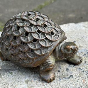 May include: Bronze turtle figurine with a detailed shell composed of coin-like shapes. The sculpture has a weathered appearance, with a greenish-brown patina. The turtle's head and legs are visible, resting on a stone surface.