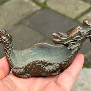 May include: A small, boat-shaped bronze dish featuring a sculpted dragon head at one end and a dragon tail at the other. The metal has a weathered, dark brown and oxidized green patina, with detailed scales visible along the sides of the vessel.