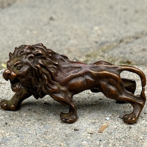 May include: A bronze lion figurine in a roaring pose. The sculpture has detailed features, including a textured mane and open mouth. The lion is depicted in a walking stance, with its tail curved upwards. The figurine is a dark brown color.