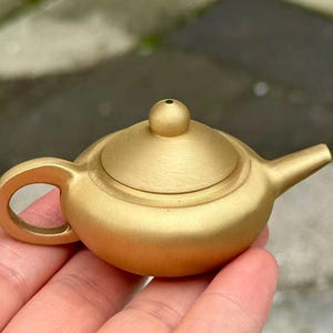 May include: A small, gold-colored teapot with a rounded body, a curved handle, and a short spout. The lid has a small knob on top. The teapot is held in a person's hand, showcasing its compact size and design.