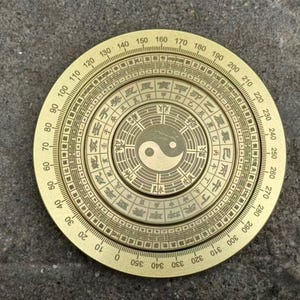Könnte beinhalten: Ein runder, messingfarbener Kompass mit aufwendigen Gravuren und chinesischen Schriftzeichen. Der Kompass hat ein zentrales Yin-und-Yang-Symbol und eine kreisförmige Skala mit Gradmarkierungen von 0 bis 360. Ein Feng-Shui-Werkzeug.