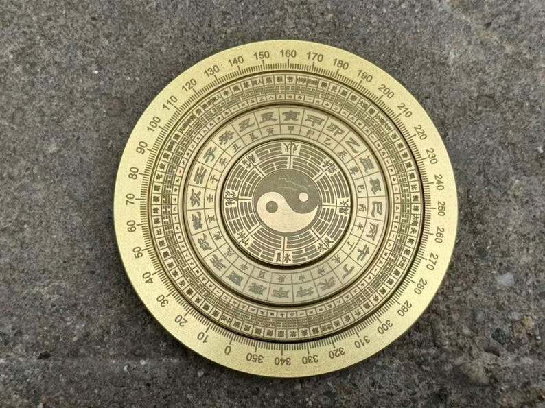May include: A round, brass-colored compass with intricate engravings and Chinese characters. The compass features a central Yin and Yang symbol and a circular scale with degree markings from 0 to 360. The compass is a tool for Feng Shui.