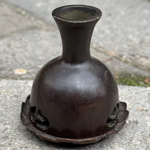 May include: A dark brown, bell-shaped metal incense burner with a narrow neck and a flared rim. The base features a wide, circular tray with a scalloped edge and decorative cutouts around the lower perimeter of the main body structure.
