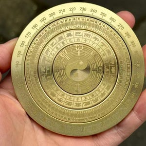 May include: A brass compass with engraved markings and a central Yin Yang symbol. The circular design features degree markings and Chinese characters. The compass is held in a hand, showcasing its size and detail.