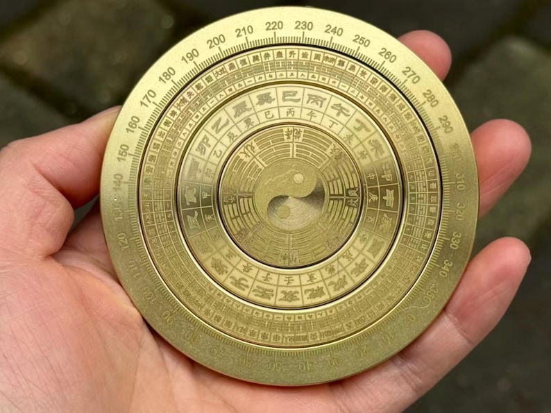 May include: A brass compass with engraved markings and a central Yin Yang symbol. The circular design features degree markings and Chinese characters. The compass is held in a hand, showcasing its size and detail.