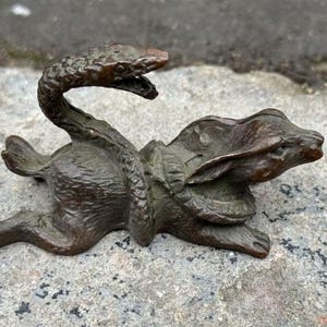 Op de afbeelding: Een bronzen sculptuur die een slang voorstelt die om een konijn gewikkeld is. De kop van de slang is omhoog en de bek is open. Het konijn ligt met zijn oren naar achteren. De sculptuur heeft een gedetailleerde textuur en een donkerbruine patina.
