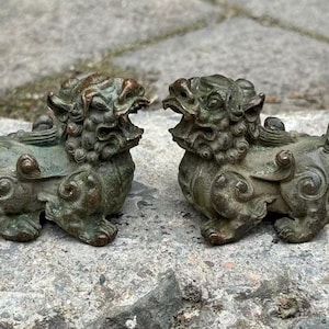 Może przedstawiać: Dwie antyczne posągi psów Foo z brązu. Każdy posąg siedzi z groźną miną, szczegółową grzywą i zakręconym ogonem. Posągi mają zwietrzałe, zielonkawe wykończenie, sugerujące wiek i wystawę na zewnątrz.