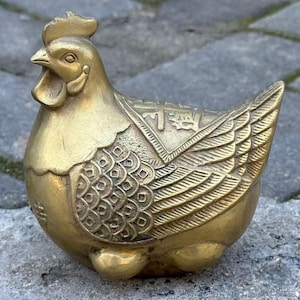 Könnte beinhalten: Eine goldfarbene, dekorative Hühnerfigur. Das Huhn befindet sich in einer sitzenden Position mit detaillierten Flügel- und Schwanzfedern. Die Figur hat einen detaillierten Kamm und Kehllappen und ist wahrscheinlich aus Messing oder Bronze.