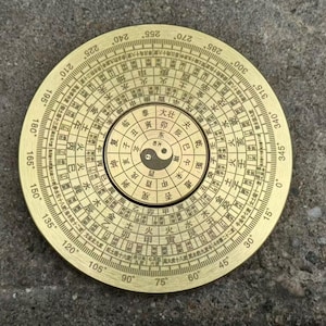 May include: A brass Feng Shui compass with detailed markings and a central Yin Yang symbol. The compass features degree markings and Chinese characters, suggesting its use in traditional practices. The circular design is set against a textured background.