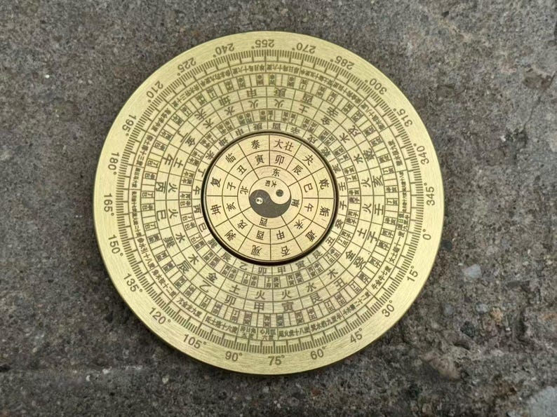 May include: A brass Feng Shui compass with detailed markings and a central Yin Yang symbol. The compass features degree markings and Chinese characters, suggesting its use in traditional practices. The circular design is set against a textured background.