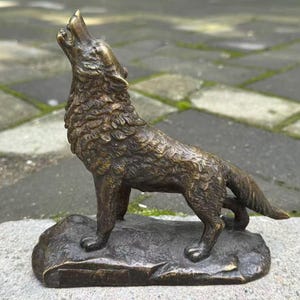 May include: A bronze-toned metal figurine of a wolf standing on a rocky base. The wolf is depicted in a howling position with its head tilted upward, showing detailed fur texture and a metallic finish throughout the entire sculpture.