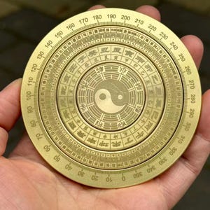 May include: A brass compass with intricate engravings and a central Yin Yang symbol. The compass features degree markings from 0 to 360 and Chinese characters. The compass is held in a hand.