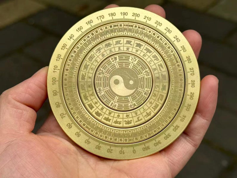 May include: A brass compass with intricate engravings and a central Yin Yang symbol. The compass features degree markings from 0 to 360 and Chinese characters. The compass is held in a hand.