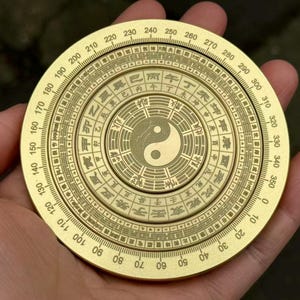 May include: A golden, circular compass held in a hand. The compass features concentric rings with numbers, symbols, and a central Yin-Yang symbol. The outer ring displays degree markings from 0 to 360.