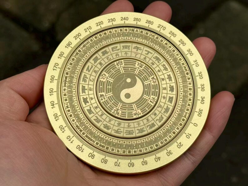 May include: A golden, circular compass held in a hand. The compass features concentric rings with numbers, symbols, and a central Yin-Yang symbol. The outer ring displays degree markings from 0 to 360.