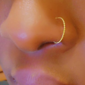Puede incluir: Primer plano de una nariz con un aro de nariz retorcido de color dorado. El aro tiene una forma curva continua, y la piel alrededor de la nariz es de un tono claro y cálido. La joya es el punto focal.