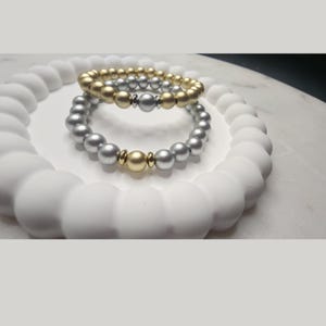 Puede incluir: Dos pulseras de cuentas, una dorada y otra plateada, descansan sobre una bandeja circular blanca texturizada. Las pulseras presentan cuentas metálicas, con detalles dorados en la pulsera plateada. Un accesorio elegante.