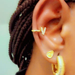 Op de afbeelding: Close-up van een gouden oor met sieraden. Een gouden oor manchet met de letter 'V' bevindt zich op het bovenste oor. Een kleine gouden oorbel en een gouden oorring zijn ook zichtbaar. De sieraden hebben een glanzende afwerking.