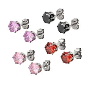 Puede incluir: Pendientes de botón en varios colores, incluyendo rosa, rojo y negro. Cada pendiente presenta una gema redonda y facetada engastada en metal plateado. Sobre un fondo blanco.