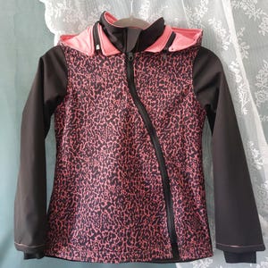 Puede incluir: Chaqueta estampada en negro y rosa con capucha. La chaqueta tiene una cremallera diagonal y mangas negras. El interior de la capucha es rosa. La chaqueta está colgada de una percha blanca.
