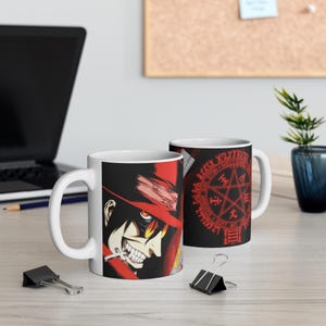 May include: A white ceramic mug featuring a graphic of a character in a red hat with yellow glasses and sharp teeth, alongside a red circular seal with a pentagram and the text Hellsing Gate Arrested Gott Mit Uns printed on a black background.