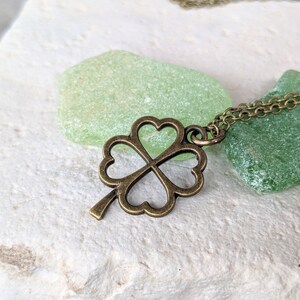 May include: A bronze four-leaf clover pendant necklace on a chain. The necklace is resting on two pieces of green sea glass.