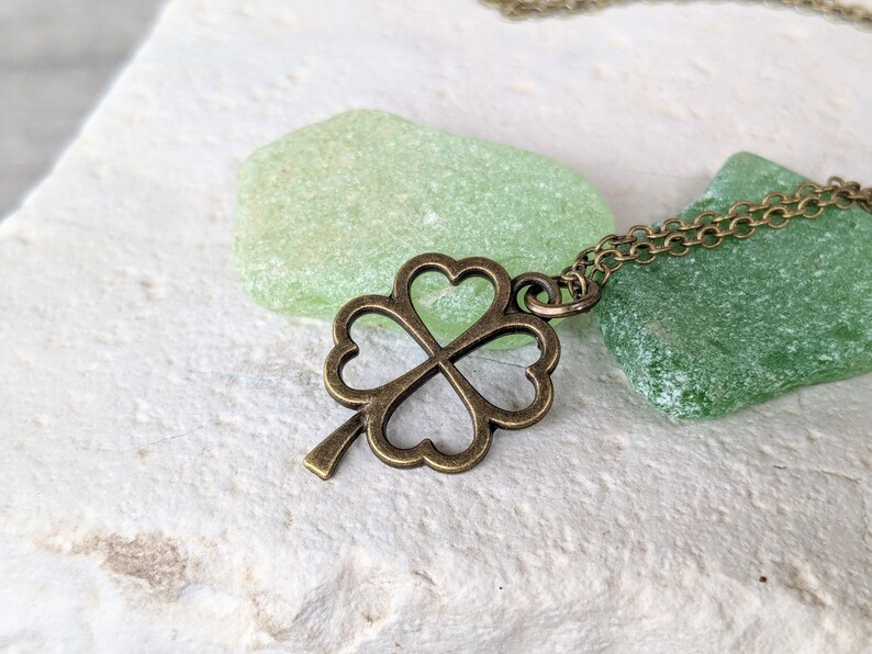 May include: A bronze four-leaf clover pendant necklace on a chain. The necklace is resting on two pieces of green sea glass.