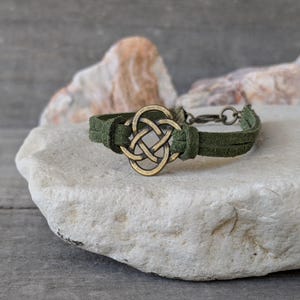 Pode incluir: Um bracelete de camur&ccedil;a verde com um amuleto de n&oacute; celta de lat&atilde;o. O bracelete est&aacute; sobre uma rocha branca.
