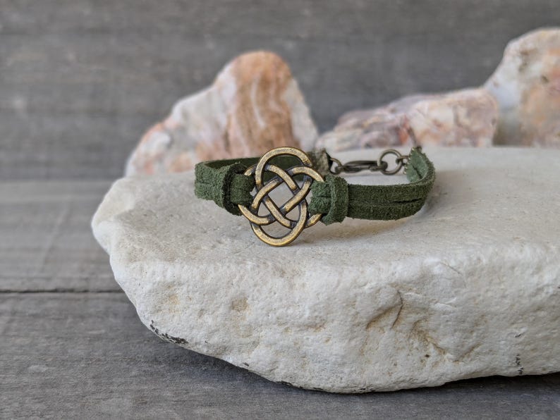 Pode incluir: Um bracelete de camur&ccedil;a verde com um amuleto de n&oacute; celta de lat&atilde;o. O bracelete est&aacute; sobre uma rocha branca.