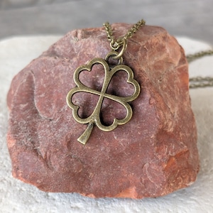 May include: A gold-toned four-leaf clover pendant necklace on a brown rock. The necklace is made of a delicate chain and the pendant is a simple, open design.