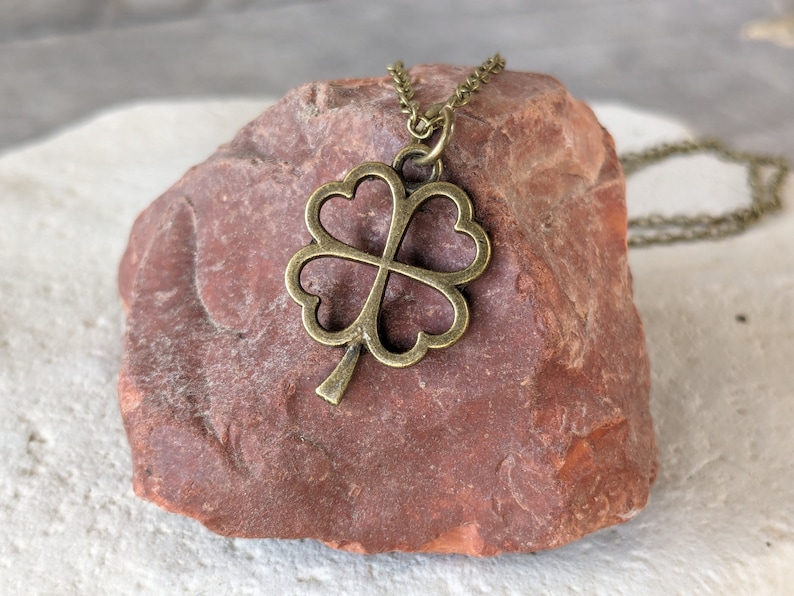 May include: A gold-toned four-leaf clover pendant necklace on a brown rock. The necklace is made of a delicate chain and the pendant is a simple, open design.