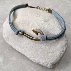 May include: A gray suede bracelet with a bronze hook charm. The bracelet is tied with a knot and has a metal clasp.
