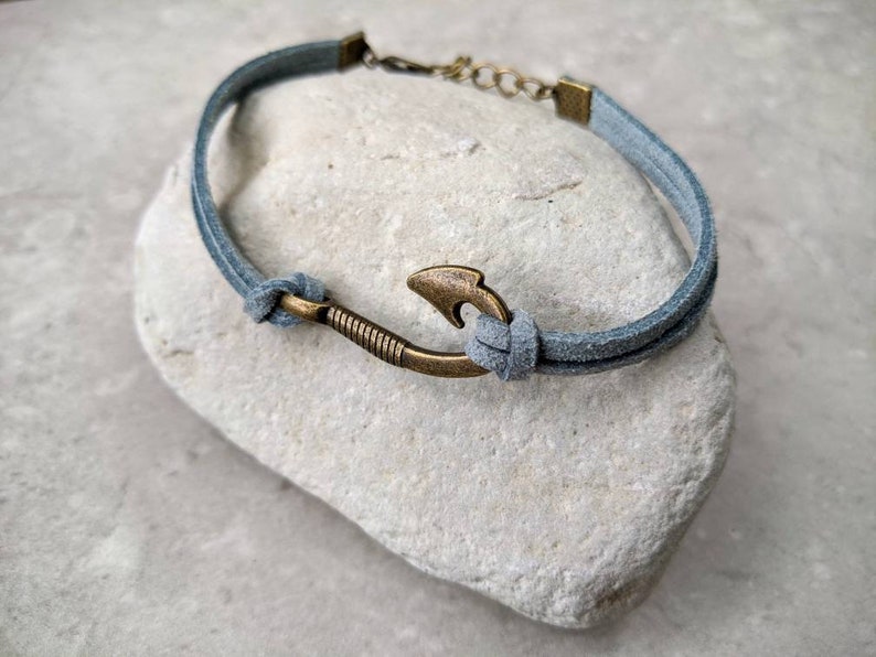 May include: A gray suede bracelet with a bronze hook charm. The bracelet is tied with a knot and has a metal clasp.