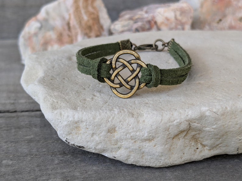 Pode incluir: Um bracelete de camur&ccedil;a verde com um fecho de n&oacute; celta dourado. O bracelete &eacute; ajust&aacute;vel e tem um fecho simples.
