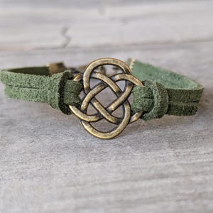 Pode incluir: Um bracelete de couro camur&ccedil;a verde com um fecho de n&oacute; celta dourado.