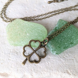 May include: A bronze four-leaf clover charm necklace with a delicate chain. The necklace is displayed on top of two green sea glass stones.