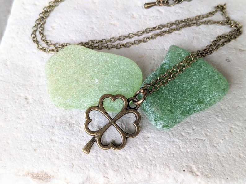 May include: A bronze four-leaf clover charm necklace with a delicate chain. The necklace is displayed on top of two green sea glass stones.