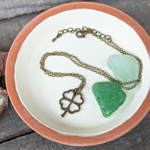May include: A gold-toned necklace with a four-leaf clover pendant, displayed on a white ceramic dish with two green sea glass pieces.