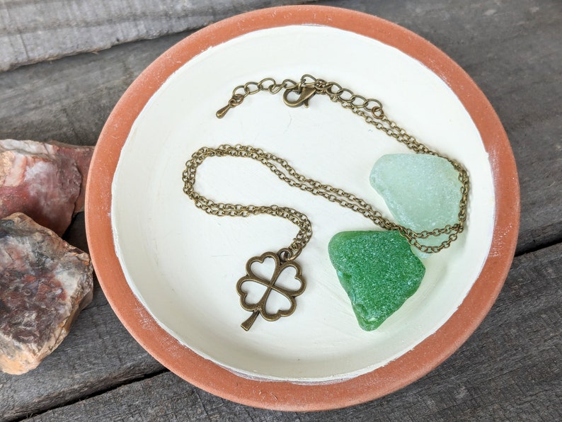 May include: A gold-toned necklace with a four-leaf clover pendant, displayed on a white ceramic dish with two green sea glass pieces.