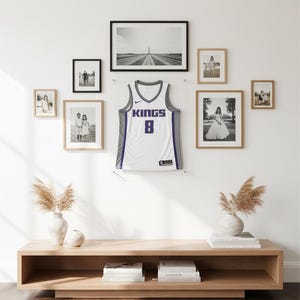 May include: A white and purple Sacramento Kings basketball jersey with the number 8 is displayed on a white wall, surrounded by framed black and white photos and family portraits. A wooden console with decorative vases and books sits below.
