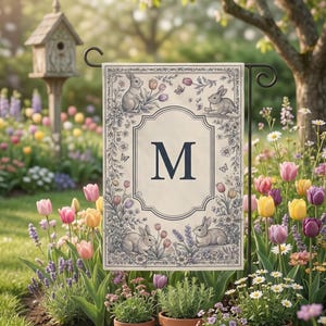 May include: A garden flag with a cream background, featuring a large navy blue letter "M" in the center. The flag is decorated with illustrations of rabbits, flowers, and butterflies. The flag is displayed in a garden with colorful tulips and other flowers.