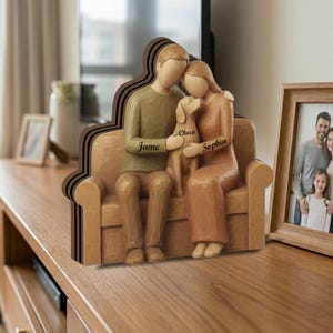 Op de afbeelding: Een houten sculptuur van een stel en een hond die op een bank zitten. De sculptuur is in bruin- en beigetinten. De namen "Jame" en "Sophia" staan op de sculptuur gegraveerd, samen met de naam van de hond, "Choco".