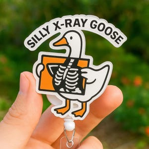 Puede incluir: Una insignia blanca y naranja con un ganso de dibujos animados sosteniendo una radiografía. El ganso tiene un esqueleto negro visible. La insignia dice "SILLY X-RAY GOOSE" en fuente blanca. La insignia está unida a un carrete retráctil.