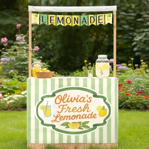 以下が含まれることがあります： 白いキャノピーと「LEMONADE」と書かれたバナーが付いたレモネードスタンド。スタンドは緑と白のストライプのスカートと「Olivia's Fresh Lemonade」と書かれた看板が特徴です。レモンのバスケットと大きなガラス瓶のレモネードが見えます。