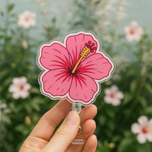May include: A pink hibiscus flower badge reel with a clear plastic retractable clip. The flower has red veins and a yellow stamen. The badge reel is held in a hand against a blurred background of flowers.
