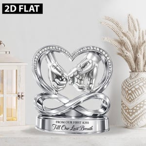 May include: A silver sculpture of two hands interlocked at the pinky fingers, forming a heart shape. The names "Anna" and "James" are inscribed. The base reads "FROM OUR FIRST KISS Till Our Last Breath."