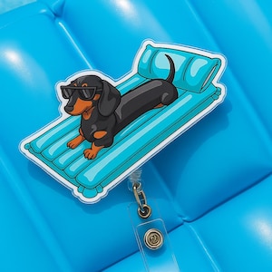 Peut inclure: Un porte-badge avec un dessin animé de teckel portant des lunettes de soleil, allongé sur un matelas gonflable bleu. Le chien est noir et marron, avec un contour blanc. Le porte-badge a un clip doré et une sangle en plastique transparent.